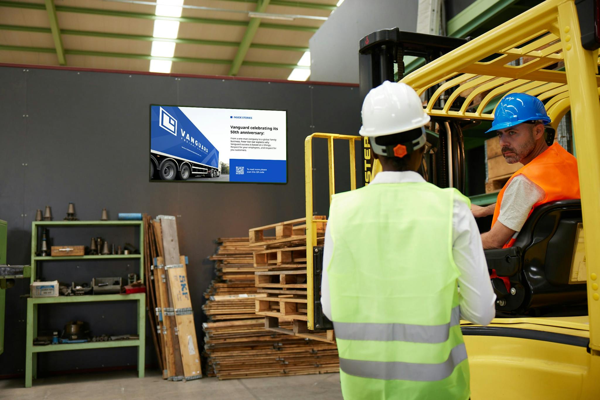 Two employees in a warehouse are speaking to each other with a digital sign in the background displaying information.